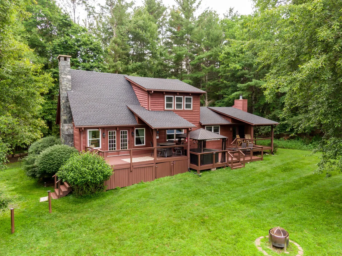A spacious wooden deck features a prominent stone chimney, a cozy fire pit area, and lush greenery all around. The space is arranged for family gatherings, outdoor meals, and relaxing by the fire under the stars, with plenty of room to play or unwind.