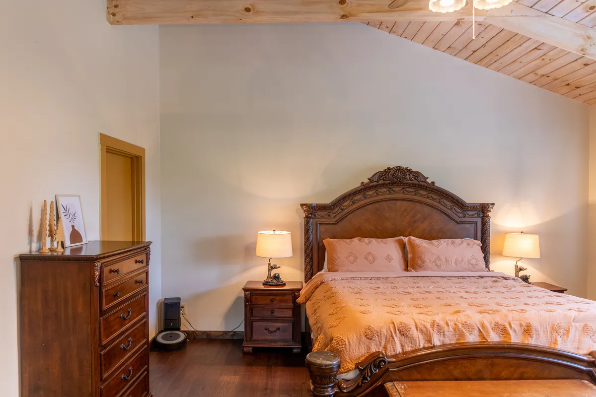 A tranquil bedroom featuring a bed with an ornate headboard, flanked by nightstands with lamps and a dresser nearby. Gentle natural light streams in, creating a restful atmosphere ideal for unwinding at the end of the day.