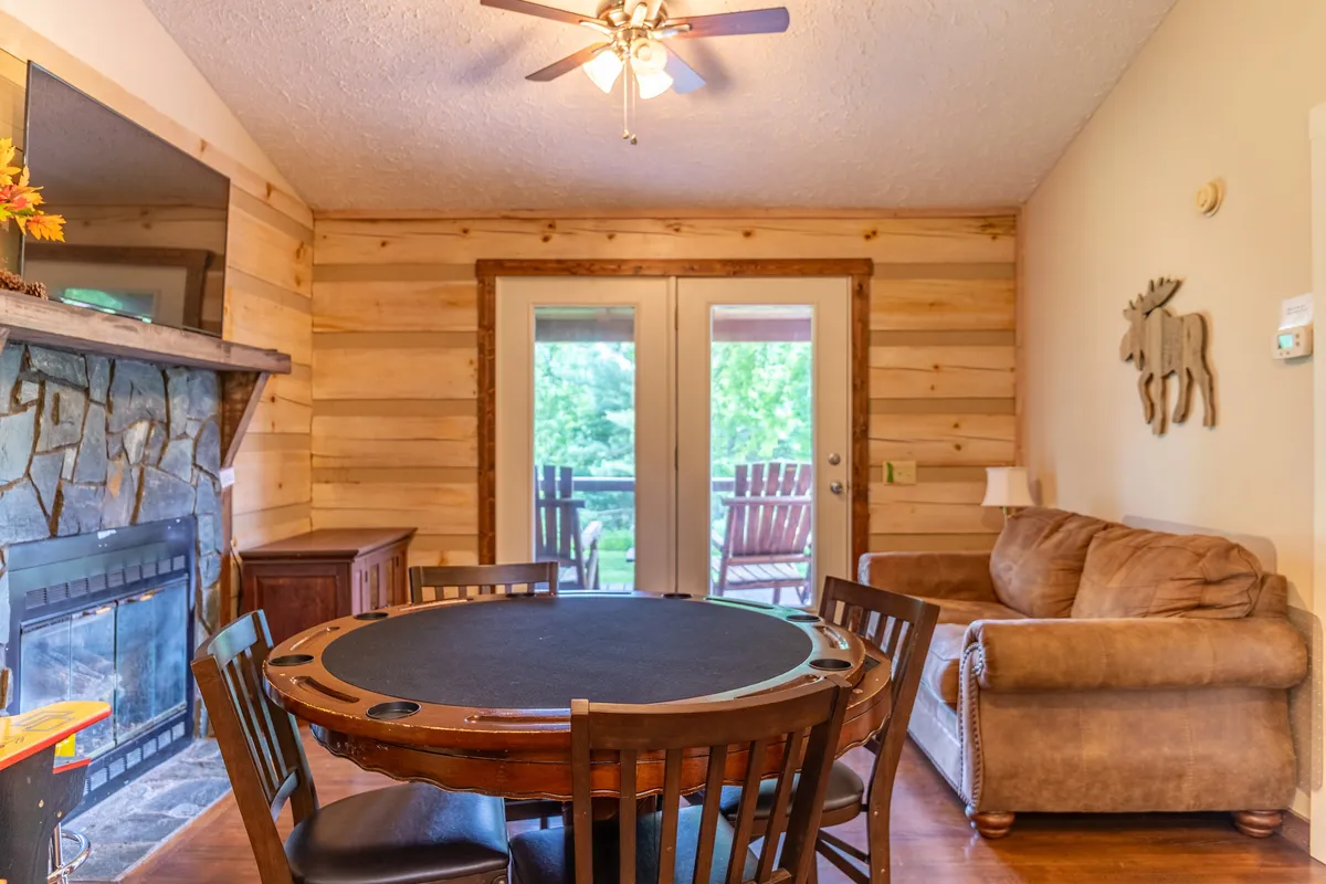 This image features a welcoming game room setup centered around a poker table in front of a cozy fireplace, with a sofa and sliding doors that open to the outdoor area. The wooden moose decoration adds a fun, playful touch, while the overall layout is ideal for gatherings, card games, or relaxing by the fire.