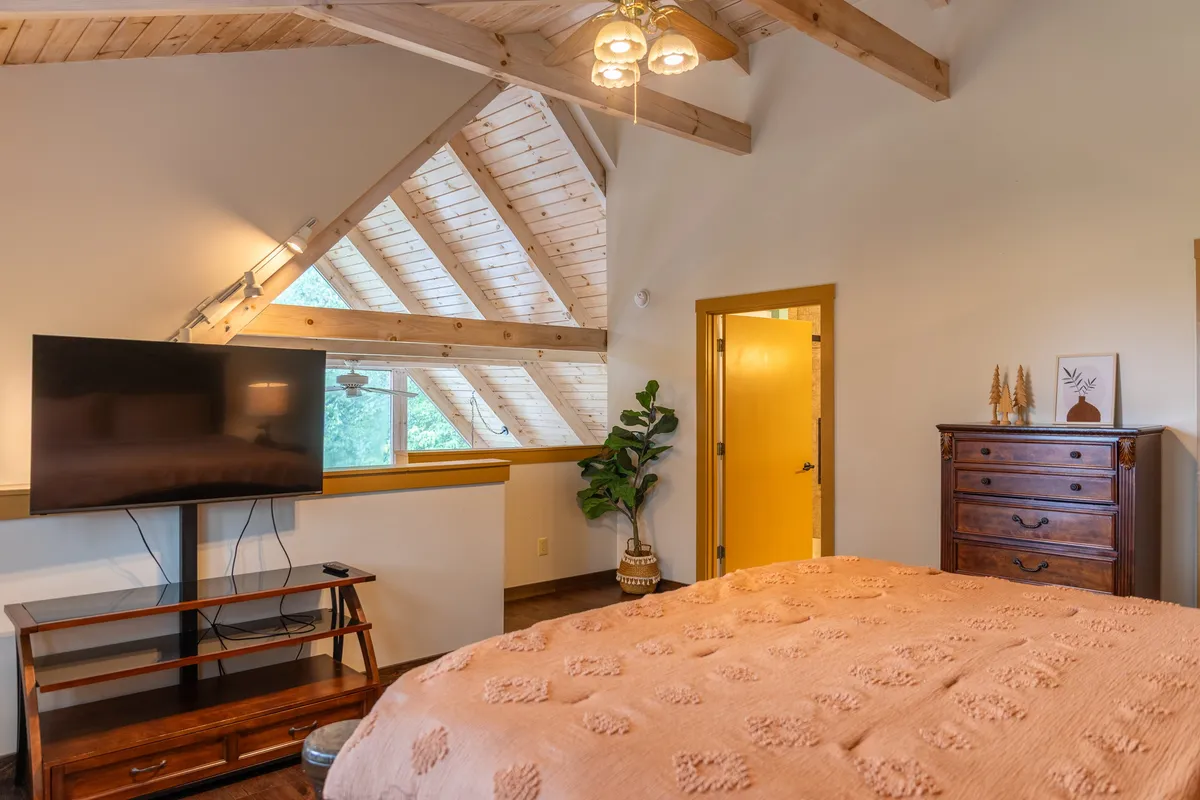 A spacious loft-style bedroom with a textured blanket on the bed, ample wooden furniture, large windows letting in natural light, an indoor plant, and a television. The space feels airy and secluded, echoing the semi-private nature of the loft master suite.