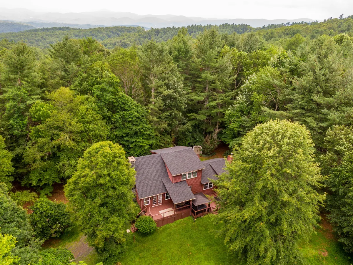 The image showcases a charming single-family home nestled amid tranquil trees, with a sweeping mountain vista stretching out in the background. A cozy porch invites relaxation, while the spacious yard promises gatherings beneath the open sky. The peaceful scene is framed by lush greenery, emphasizing the secluded, nature-immersed setting.