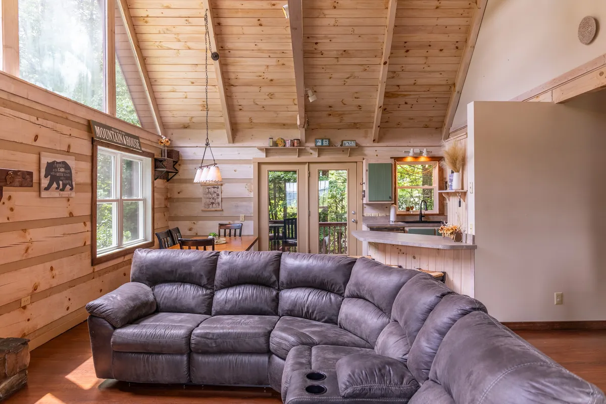 A spacious, open-concept living area seamlessly links a large sectional sofa, a wooden dining table, and a kitchen area with green cabinetry. The setting is flooded with natural light and designed for shared experiences, with ample space for lounging, socializing, and meal preparation.