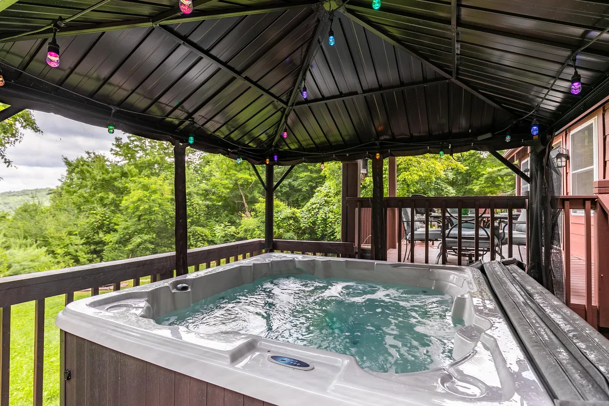 A covered outdoor hot tub surrounded by decorative string lights, with lush greenery and distant mountain views in the background. The scene is tranquil and set up for group relaxation, with guests enjoying the warm water and the inviting ambiance.
