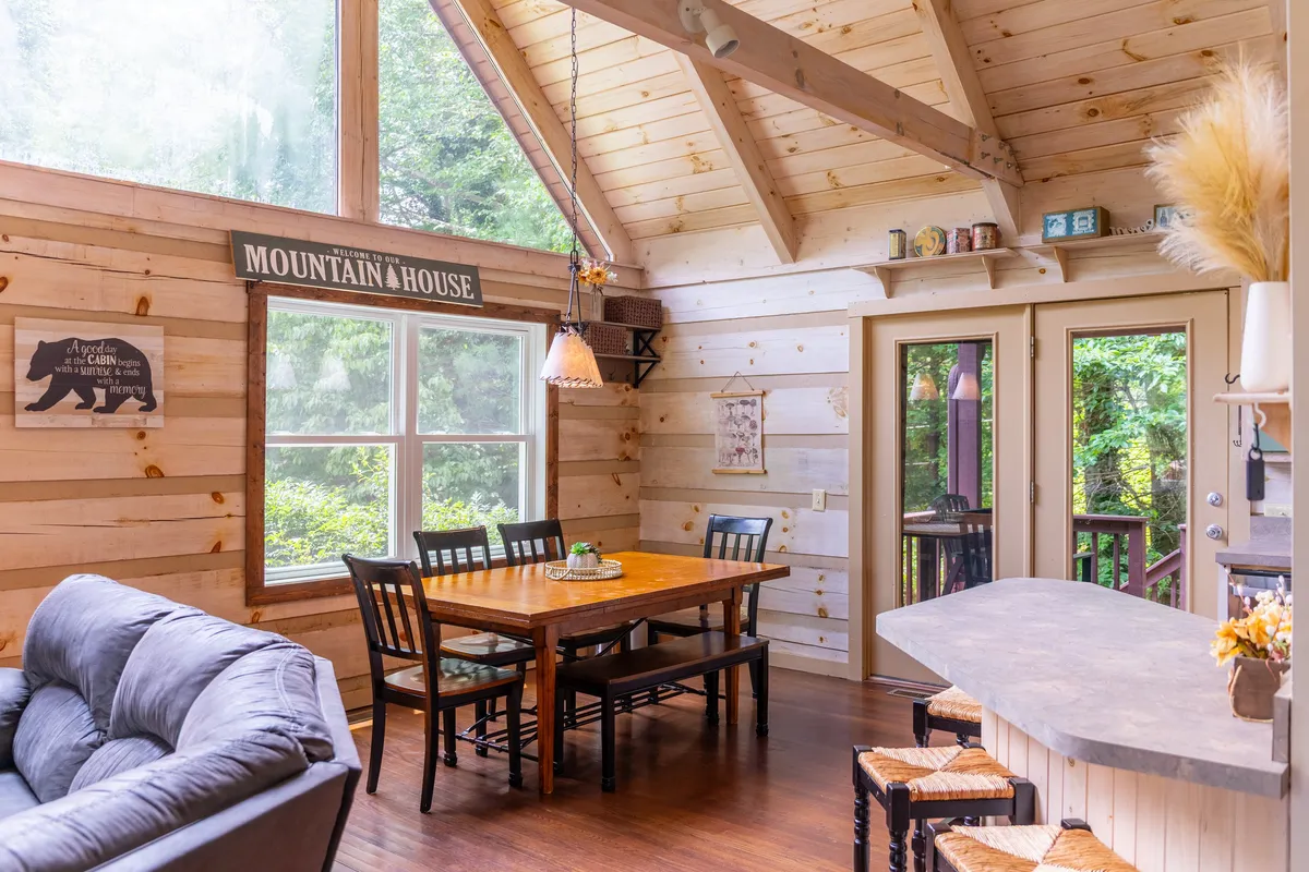 A living and dining space with large windows pouring in natural light, a rustic wooden dining table, and a plush, comfortable sofa. The ambiance is warm and inviting, ideal for gatherings and meals with loved ones while enjoying views of the outdoors.