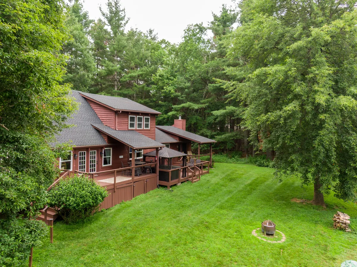A view of the home's wooden deck, adjacent fire pit, green grass, and surrounding trees. The setting is peaceful, emphasizing opportunities for outdoor family gatherings and activities like storytelling around the fire pit or roasting s'mores under the stars.