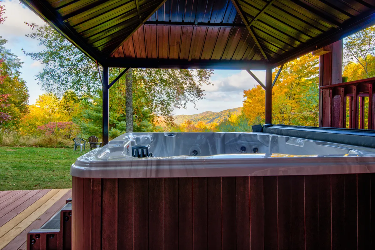 A covered hot tub sits on a wooden deck with a gazebo structure, surrounded by autumn foliage and offering a panoramic mountain view. The tranquil setting is perfect for relaxing, socializing, and stargazing, with the deck seamlessly blending into the natural landscape.